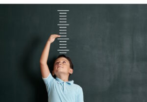 Child standing against horizontal lines drawn on blackboard to measure his height, while holding one hand above his head indicating his desired future growth
