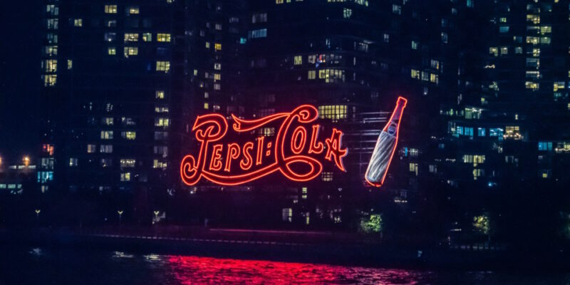 Old Pepsi-Cola neon sign lit up at night with buildings in background